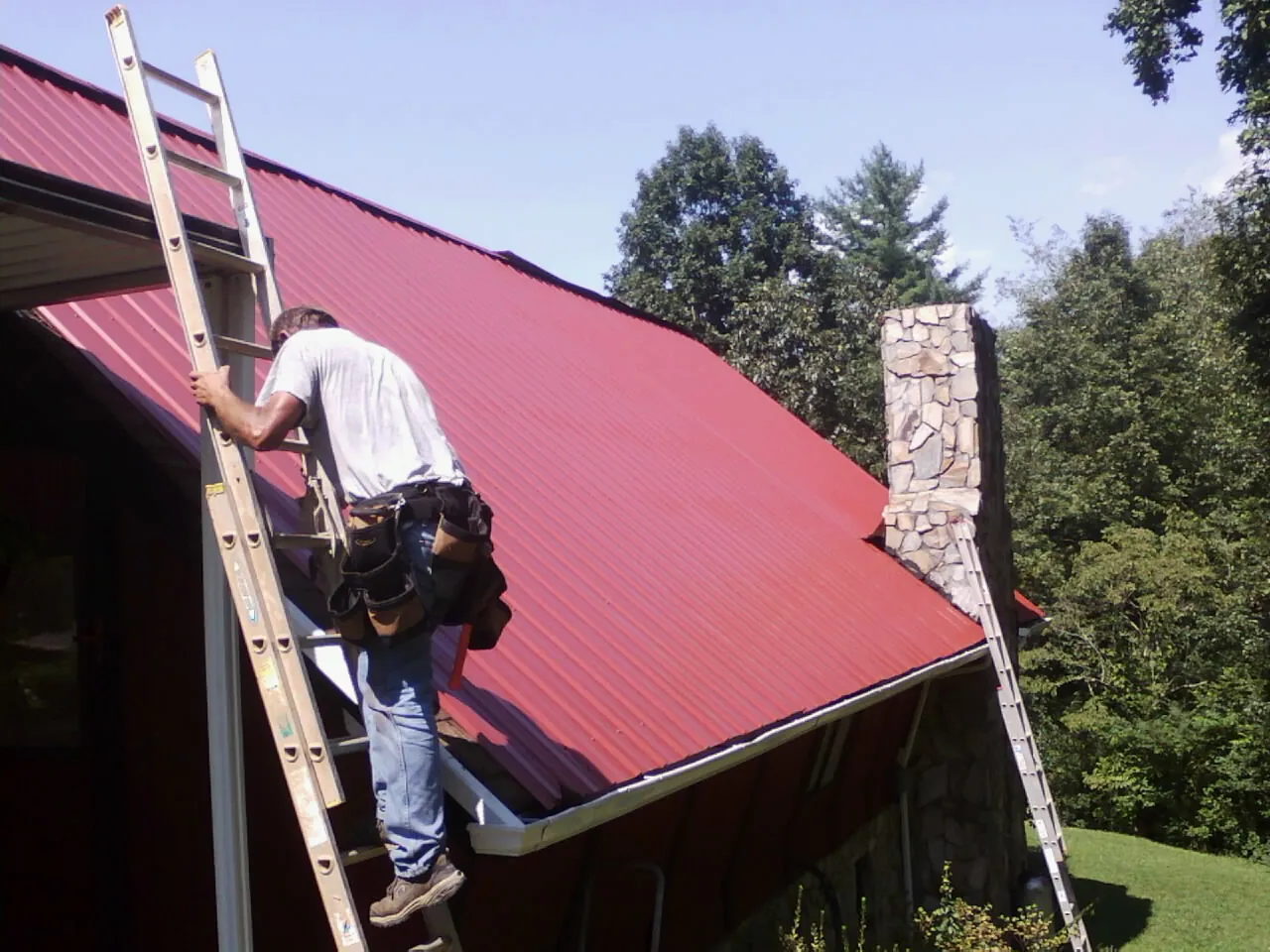 Roofer on a ladder beside a red metal roof and stone chimney — Soffit & Fascia Repair by Roofing Brothers in Meeker